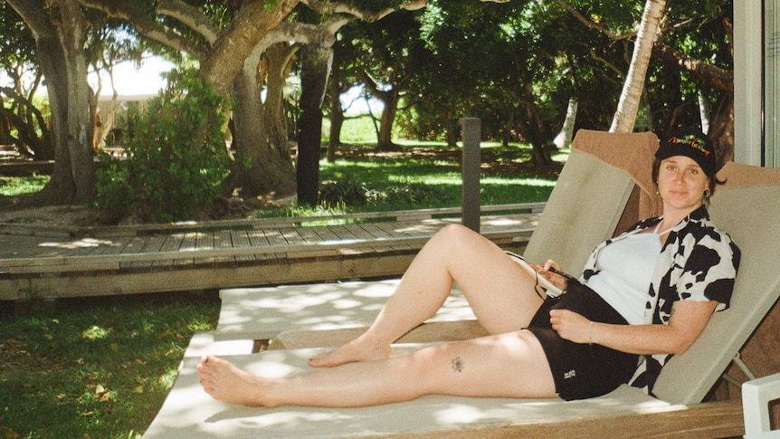 a woman wearing black shorts, white singlet and black and white shirt is lieing on a deck chair surrounded by greenary