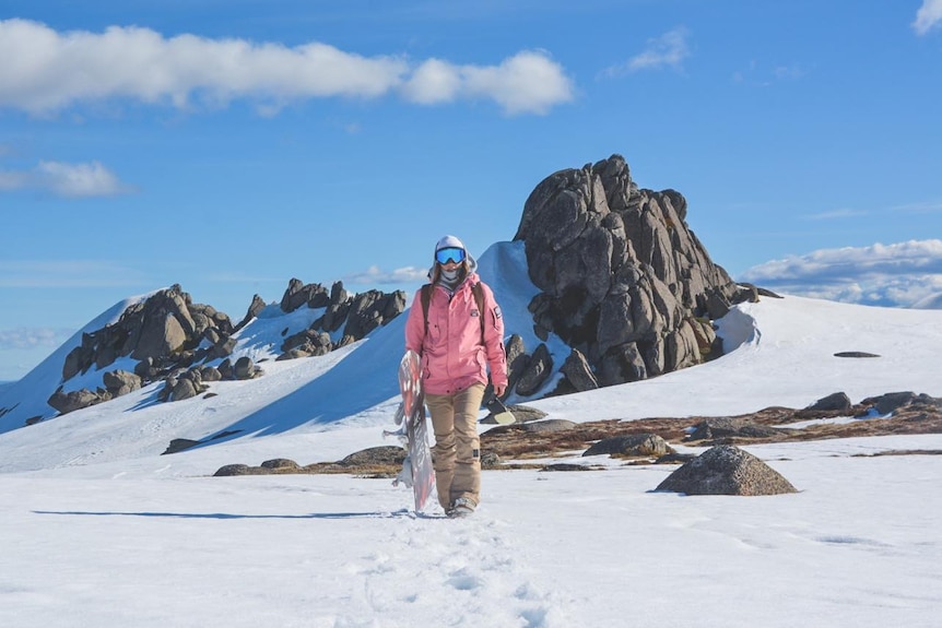 Rani is seen walking towards the camera with her snowboard standing on top of a mountain with snow and rocks behind her.