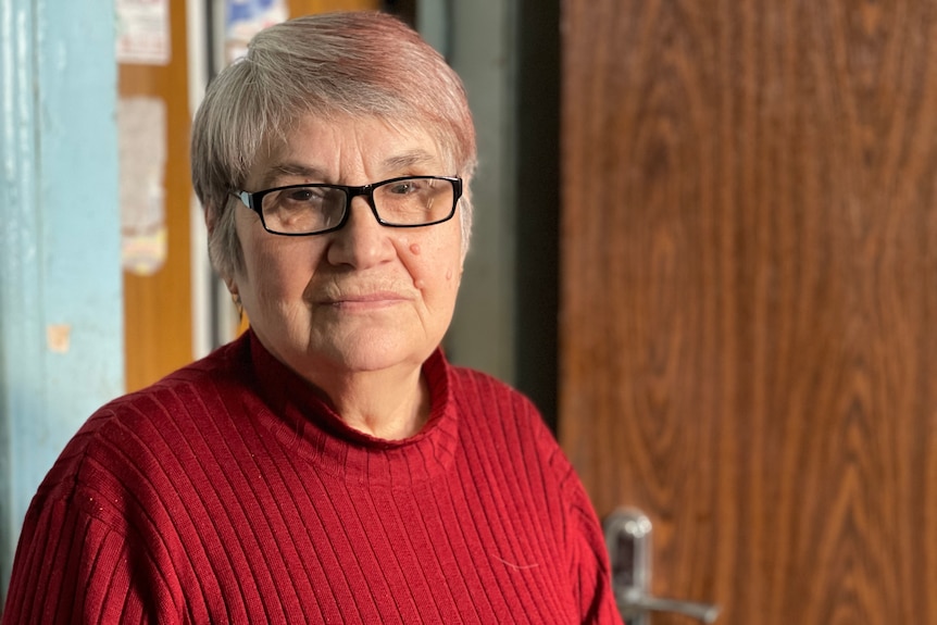 An older woman in glasses and a red sweater stands solemnly.