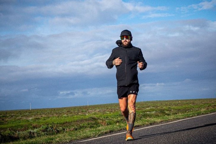 A man in black clothing running on a road with green fields and blue skies in the background