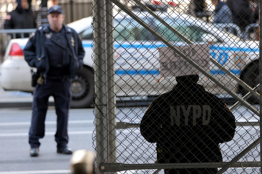 Men in uniforms that say NYPD stand next to a fence near a police car