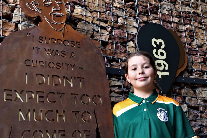 A girl wears a green school uniform and stands next to a brass cut out of her great uncle that has a quote from him on it.