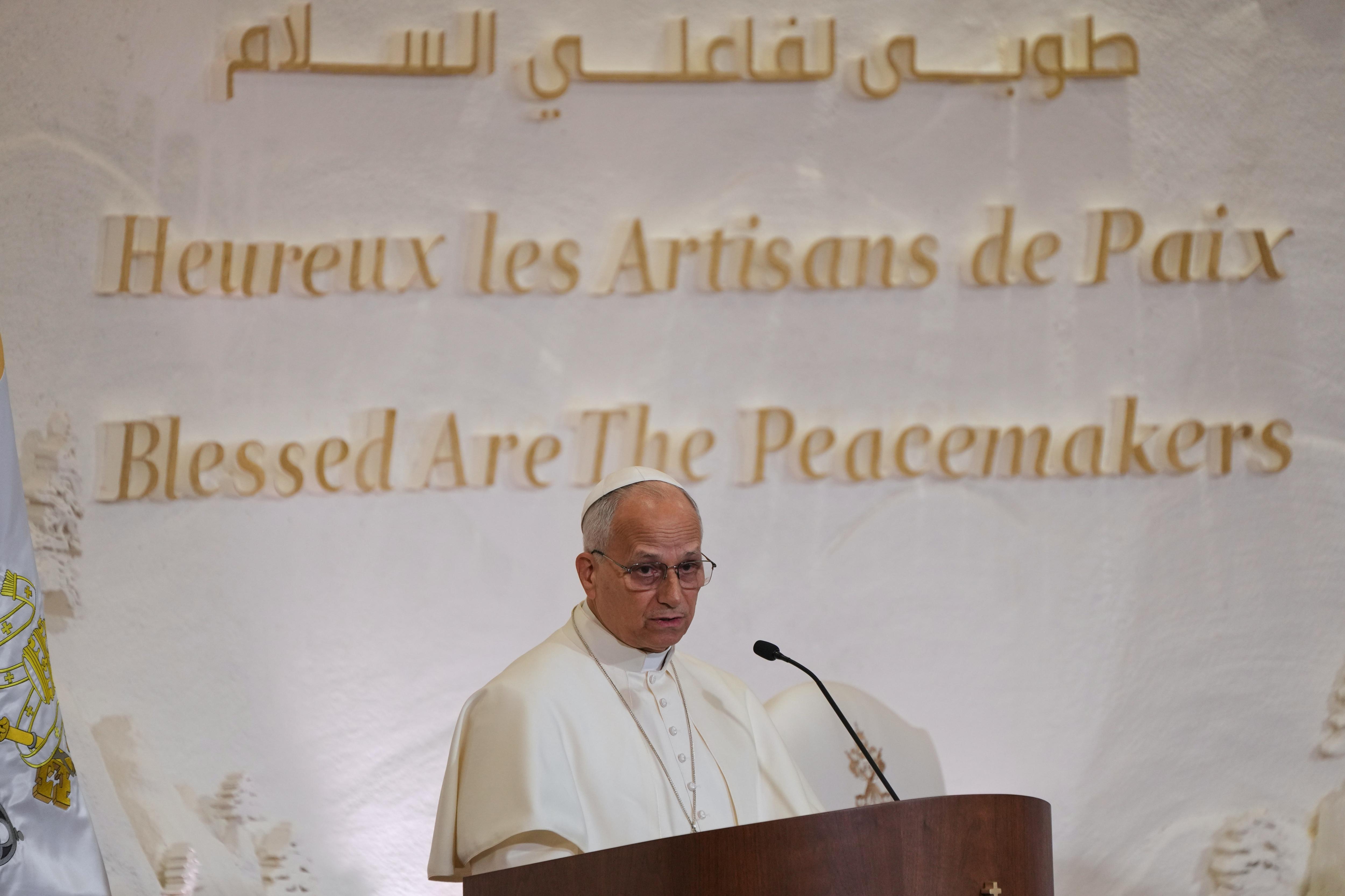 Pope Leo dressed in white standing at a podium, behind him a wall with text reading Blessed are the peacemakers
