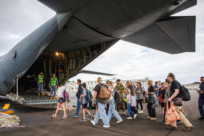 A large aeroplane sits with its back door open on the tarmac as people with bags walk toward it