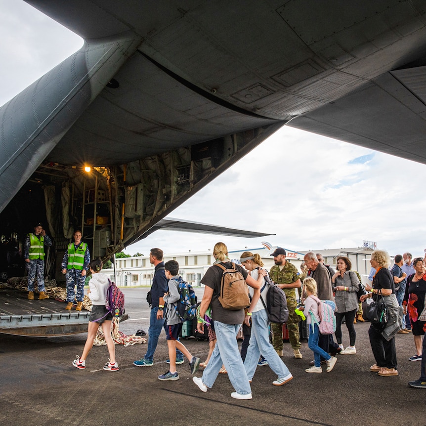 A large aeroplane sits with its back door open on the tarmac as people with bags walk toward it