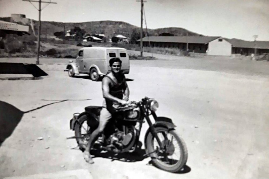 Old black and white photo of a young man sitting on a motor bike smiling for a picture 