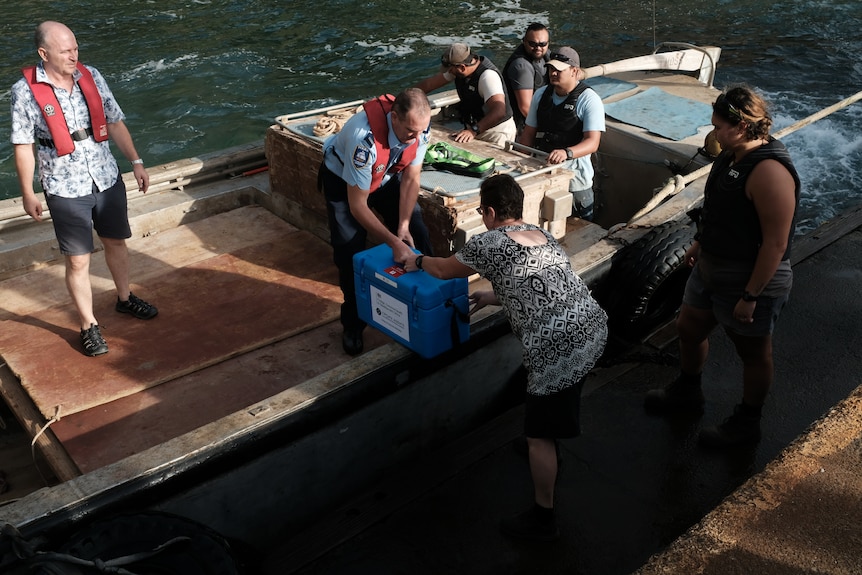 O cutie care transportă vaccinuri este transmisă din barcă către o femeie de pe doc, în timp ce oamenii stau în jurul ei. 