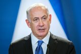 A close up photo of Benjamin Netanyahu looking off camera while speaking with a blue and white background.