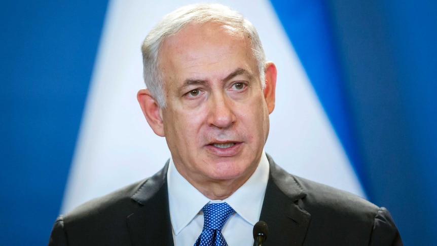 A close up photo of Benjamin Netanyahu looking off camera while speaking with a blue and white background.
