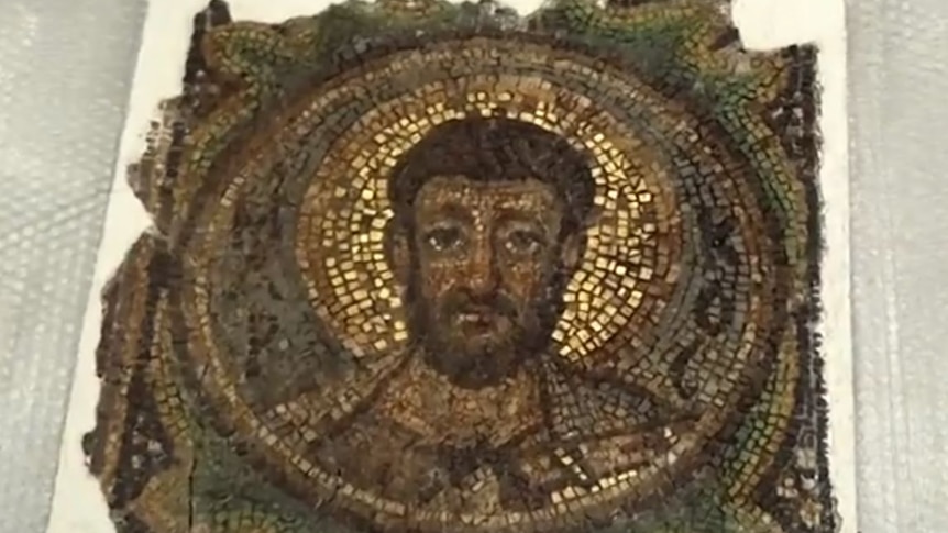 An aged mosaic depicting a young man with dark hair. The tiles look to be aged and are laid against a white backing board.