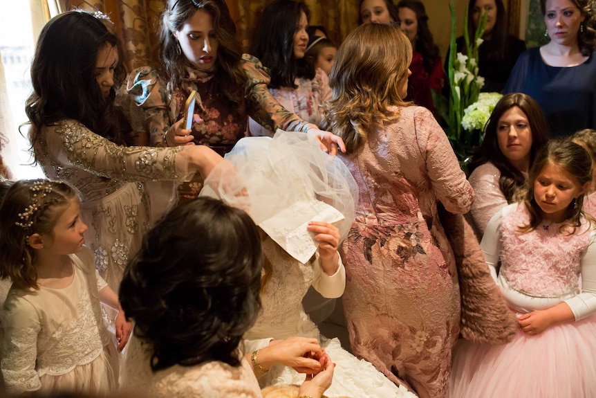 A bride sits with a white veil over her head, in the centre of a cluster of girls and women, all finely dressed.