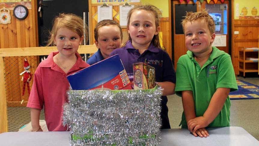 Four kindy children, two boys and two girls, stand behind a Christmas hamper full of food items.