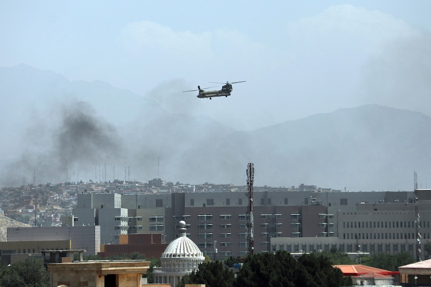 A Chinook helicopter flies low over buildings in Kabul. Smoke rises from one part of the city.