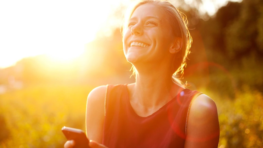 A young woman smiles and uses her phone as the sun beams down on her.