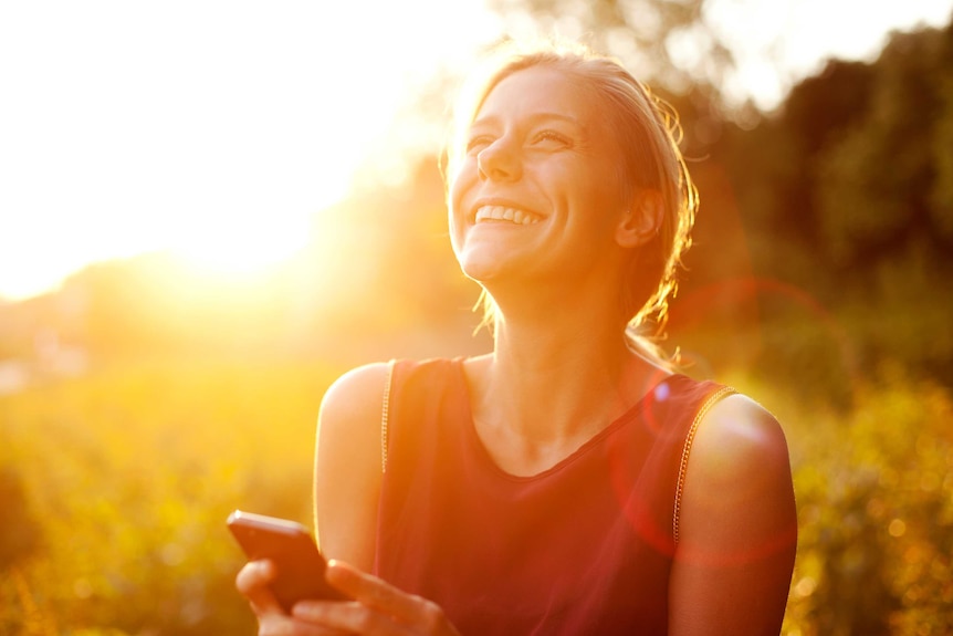 A young woman smiles and uses her phone as the sun beams down on her.