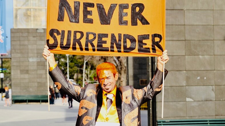 Sebastian Dell'Orefice, dressed in an orange-lettered suit holds a sign reading 'We Will Never Surrender' outside the MCG.