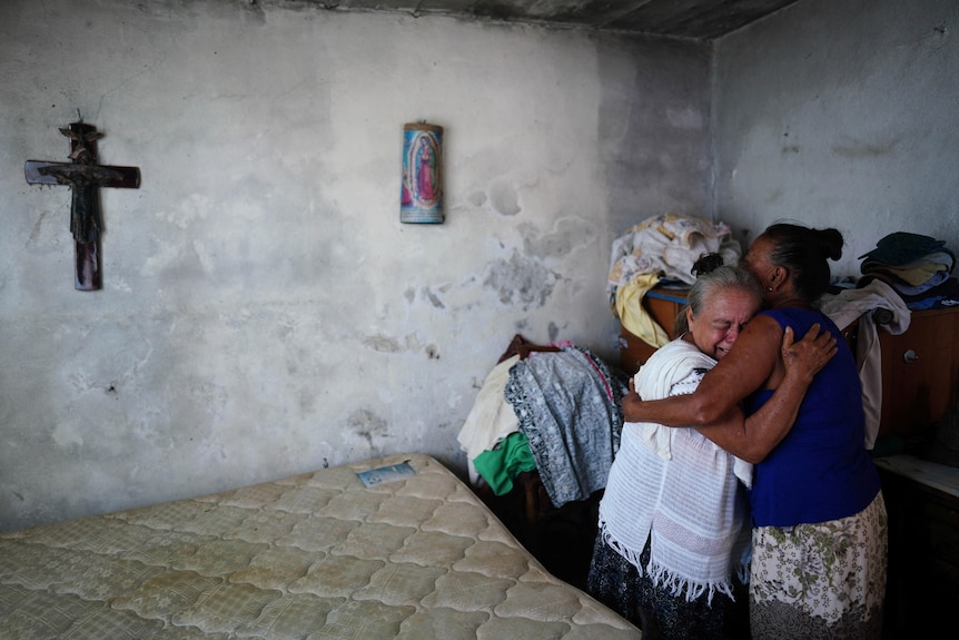 Two elderly women clutch each other as they cry inside a simple house.