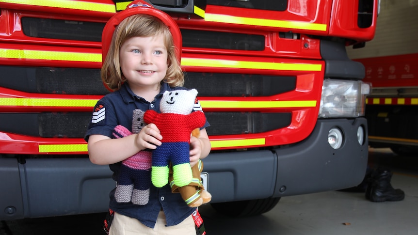 Liam Moffitt dressed in bright red helmet, custom-made firefighting uniform, holding trauma teddy 