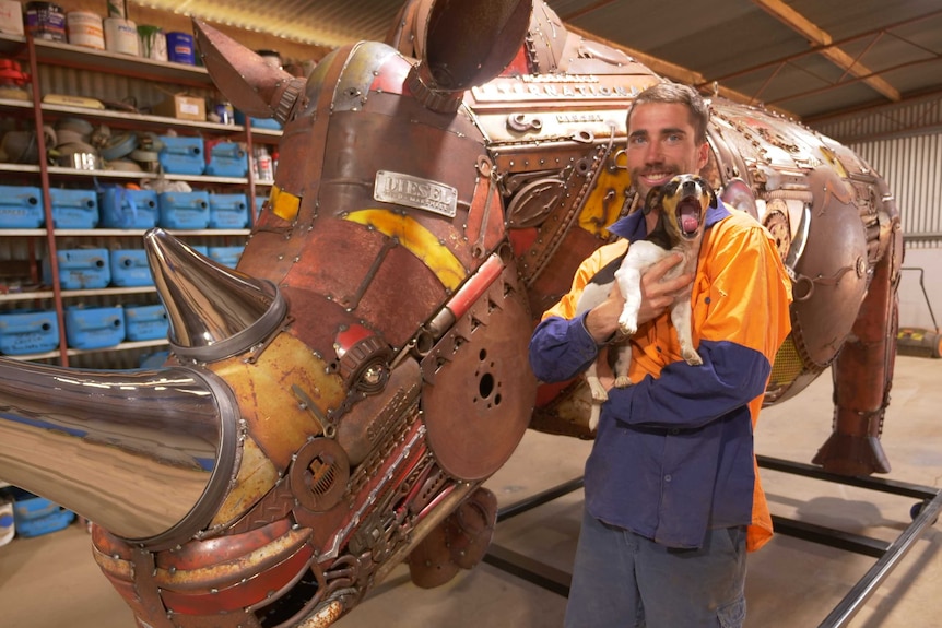 A man stands, holding a dog and smiling, in front of a huge metal rhinoceros.