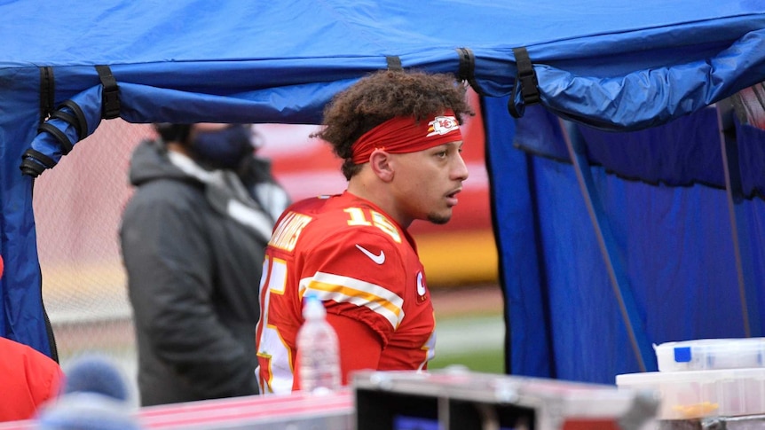 Patrick Mahomes in uniform walks into a blue tent wearing a headband