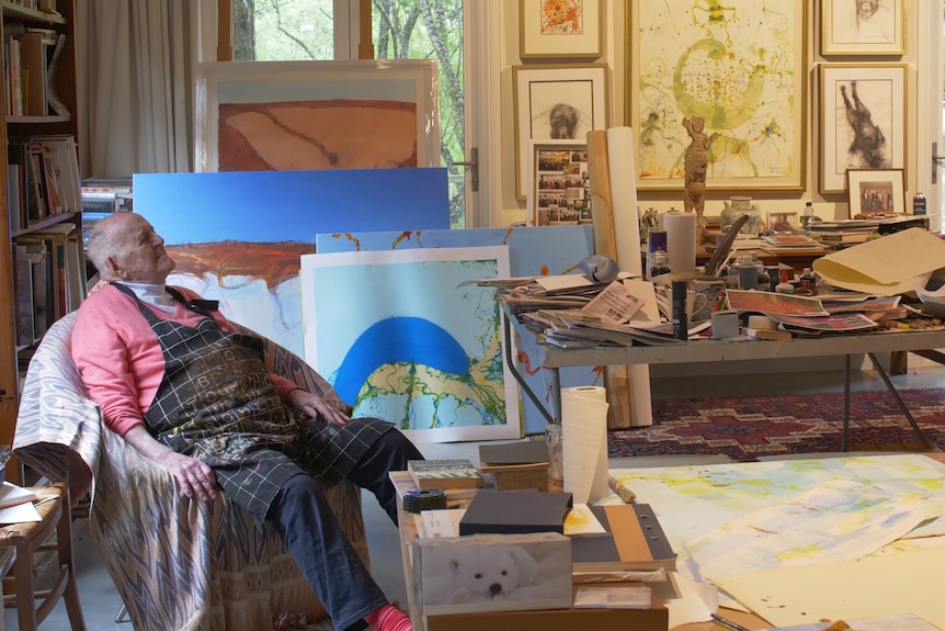 An elderly man in sweater and art smock sits in an art studio filled with canvases and tools