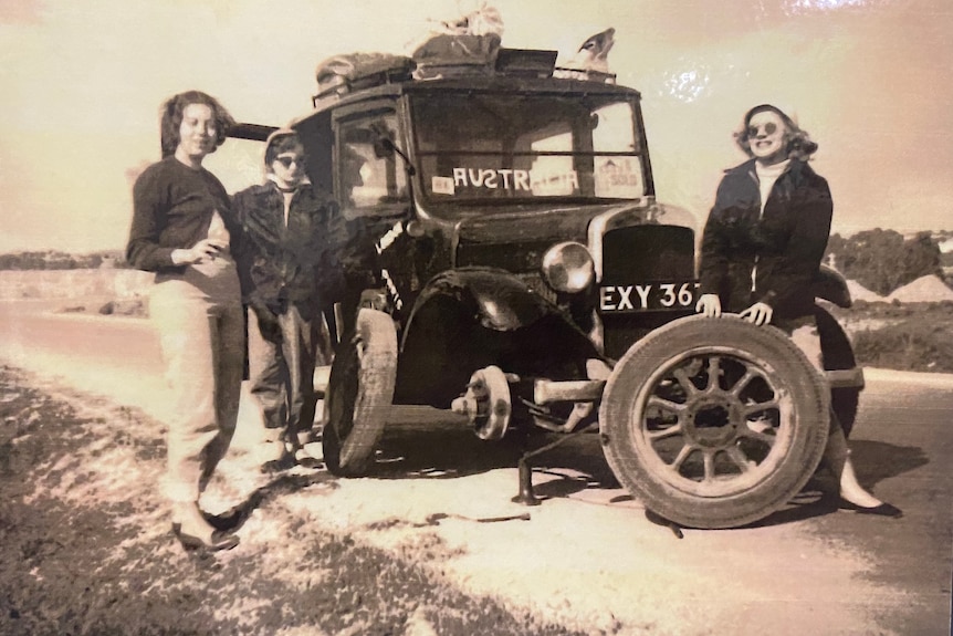 Three women stand around a black taxi with a missing wheel in a black and white photo.