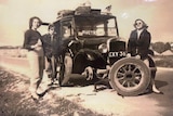 Three women stand around a black taxi with a missing wheel in a black and white photo.