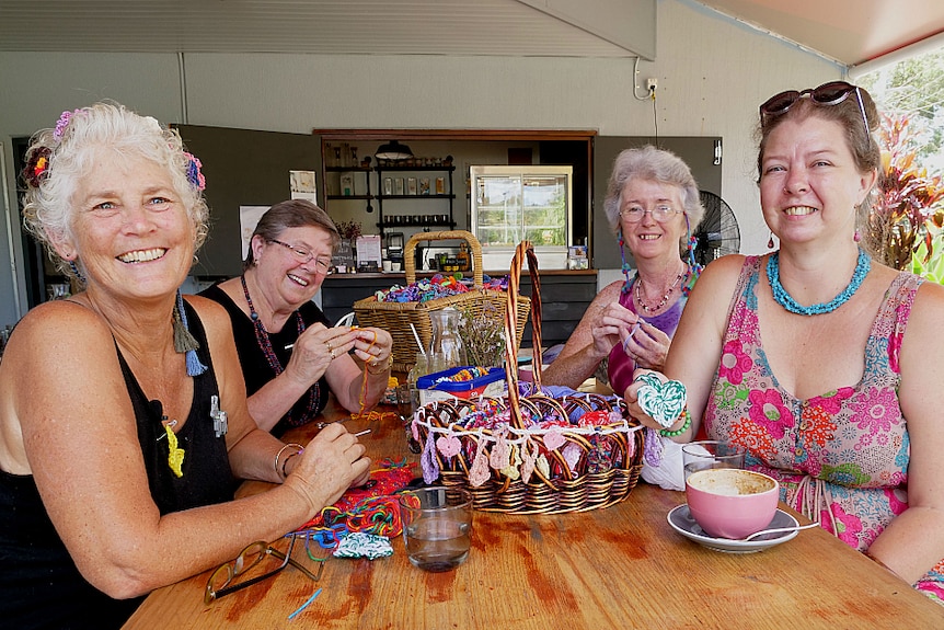 Four women sit outside at a cafe in summer, bright clothing while they crochet small hearts.