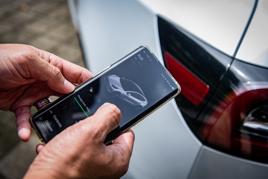 A close-up of Marc looking at an iPhone in front of his Tesla, the app says it's fully charged.