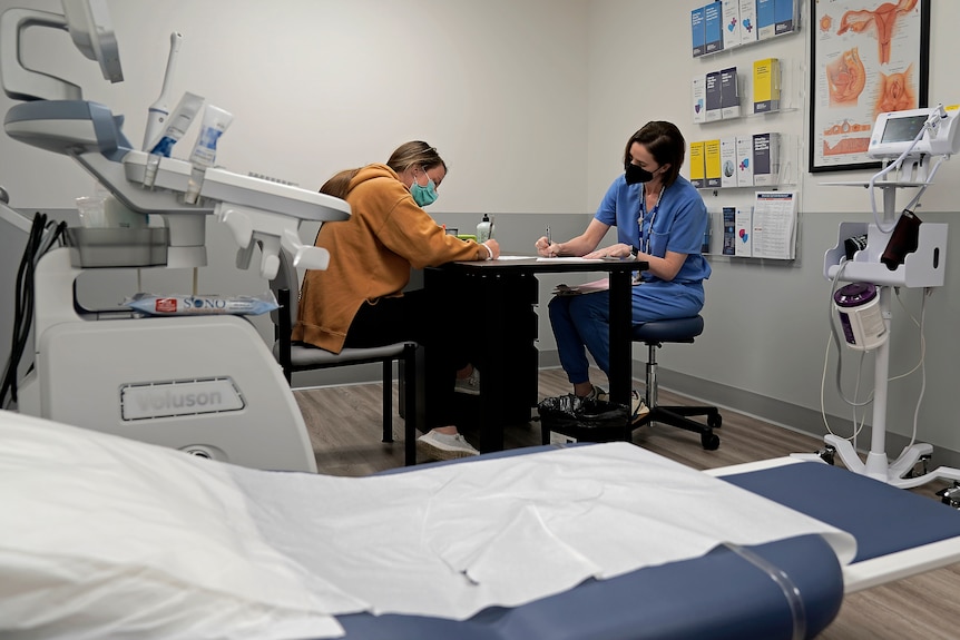 A woman and a doctor sit at a table in a dr's office next to a treatment table. 