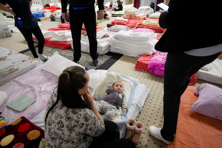 A woman sits with her infant son among stacks of folded blankets at shelter set up in a hotel.