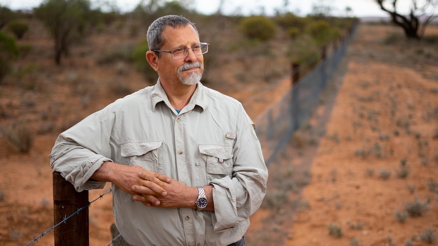 A man in a light grey button up shirt wearing glasses leans against a fence in front of red earth with trees in the background