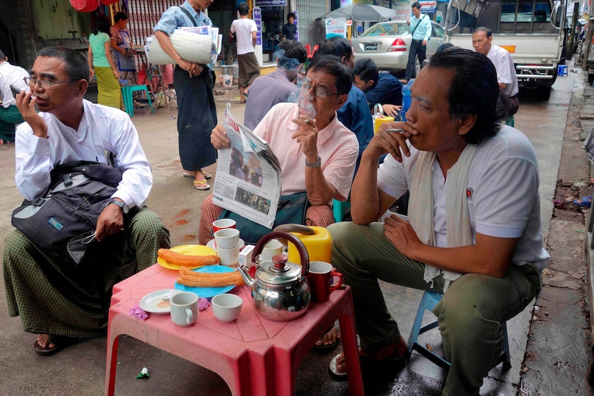 Three men sit at plastic chairs on the street, two smoke and one reads the newspaper.