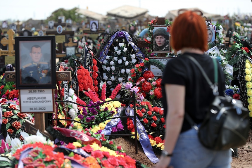 A woman at a Russian cemetery looks at the graves of Russian soldiers. They are covered in flowers. 