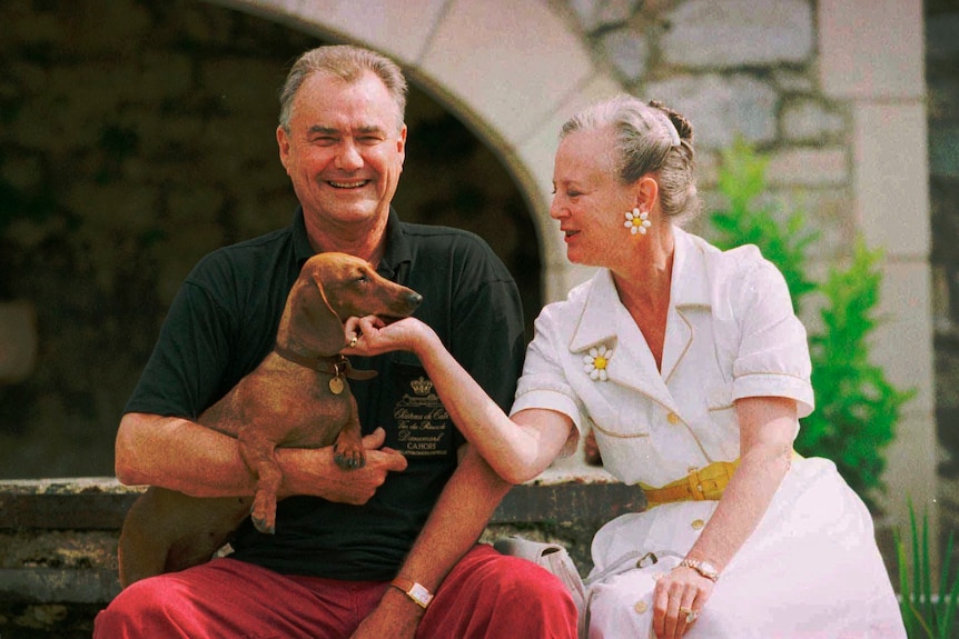 Queen Margrethe wearing daisy jewellery leans over to scratch a daschund on the chin, it sits on Henrik's lap
