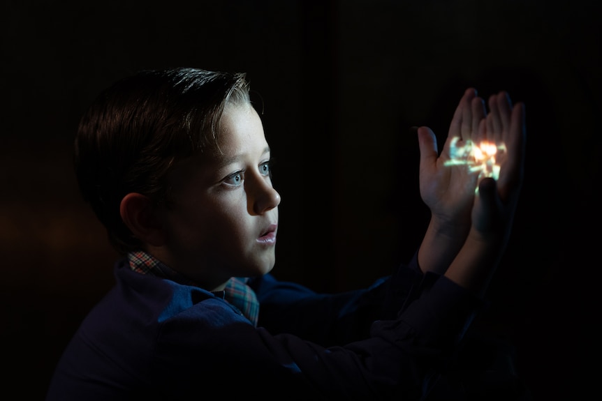 A young white boy sits in a dark room with his hands and face illuminated by a small light source.