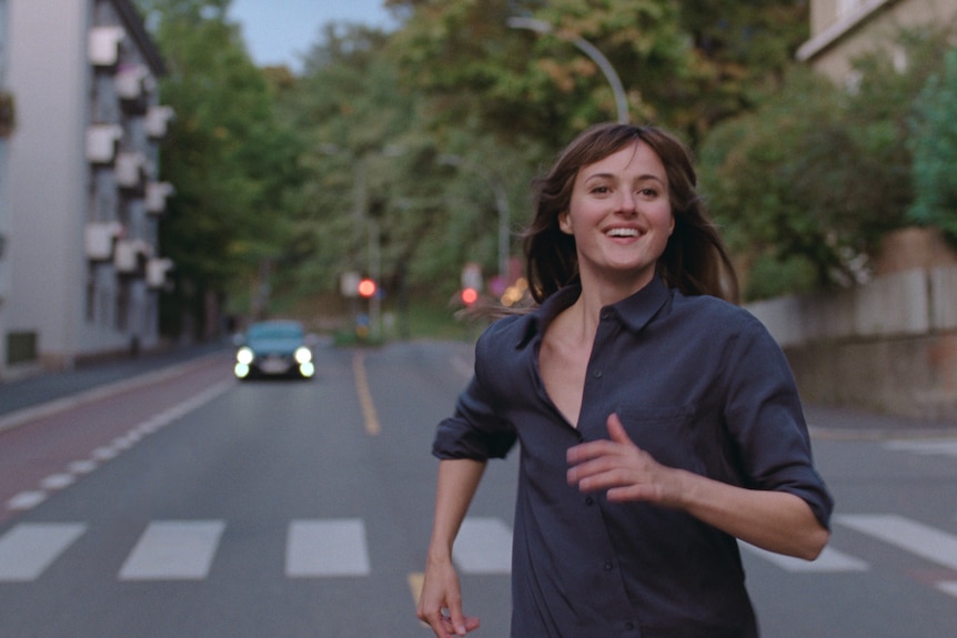 A young brunette woman in a grey shirt runs down a European street