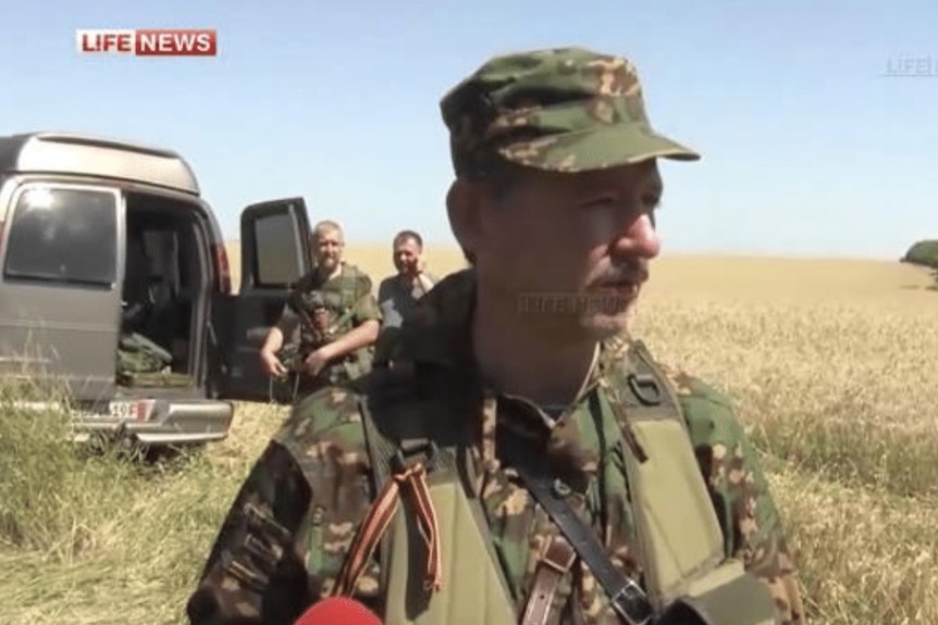 A man in military fatigues standing in a field, with two man with guns standing in front of a van in the background.