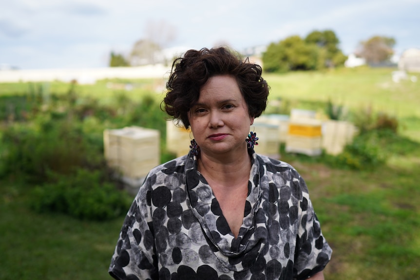 Woman wearing a black and white top stands in a field with beehives behind her.