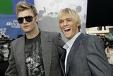 Two blond guys wearing grey jackets pose next to each other, Nick on the left is wearing big sunglasses while Aaron smiles wide