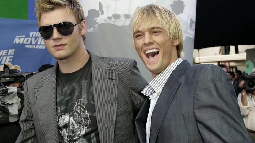 Two blond guys wearing grey jackets pose next to each other, Nick on the left is wearing big sunglasses while Aaron smiles wide