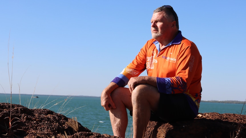 A man in an orange shirt sits on a rock and looks into the distance. The ocean is in the background. 