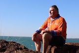 A man in an orange shirt sits on a rock and looks into the distance. The ocean is in the background. 