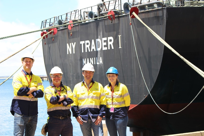 Four people wearing high-visibility shirts and hard hats standing in front of a cargo ship.