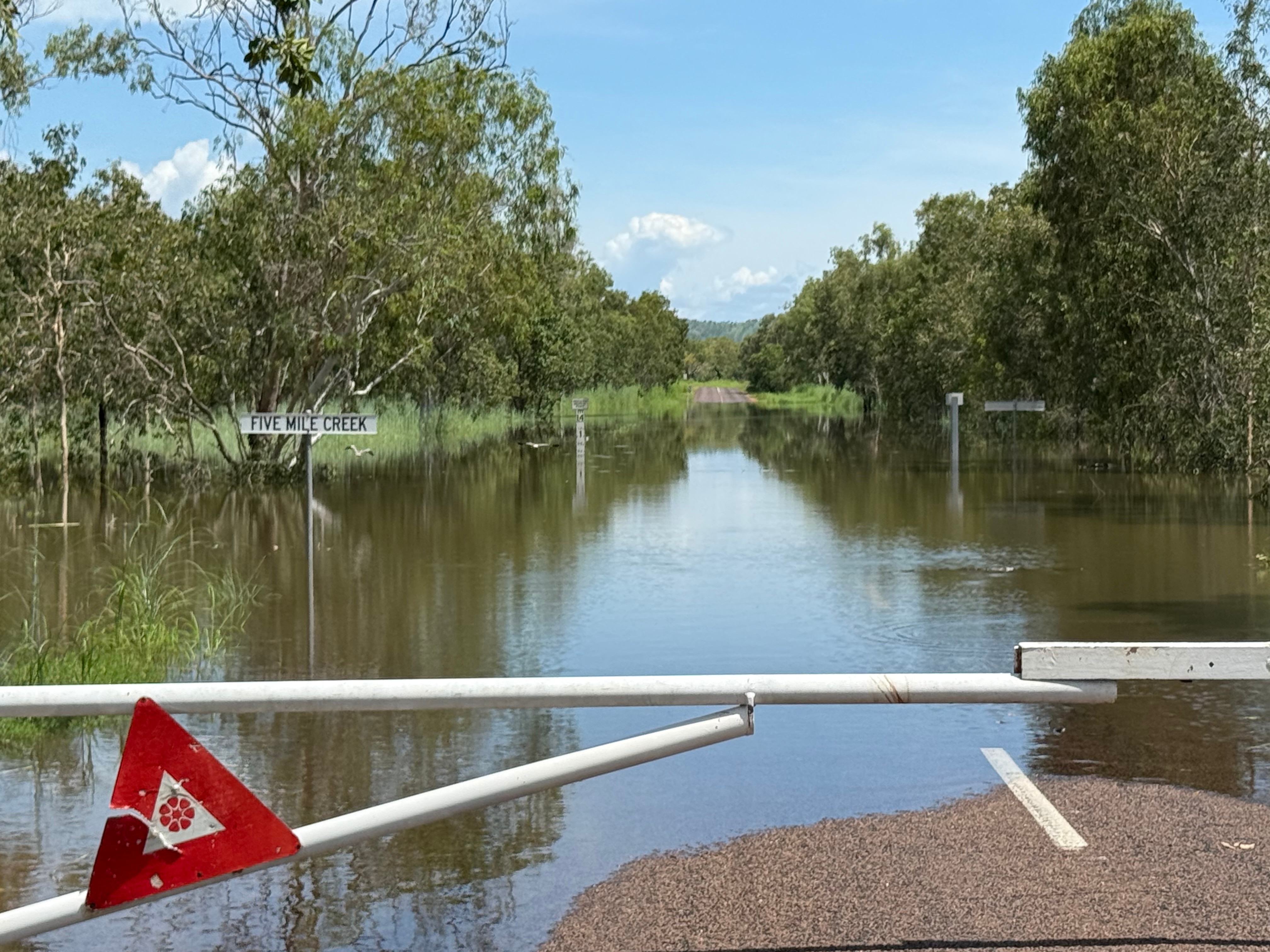 A road with white pole blocking it, water flooding a majority of the road, sign under water reads 'five mile creek'