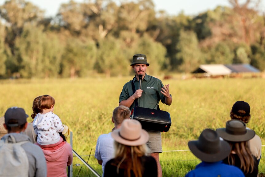 Man in green hat and shirt holding microphone speaks to group of people at a farm, with pastures in background