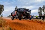 A black four-wheel-drive in the air, about a metre off the ground, with a red-dirt track beneath it and spectators watching on.