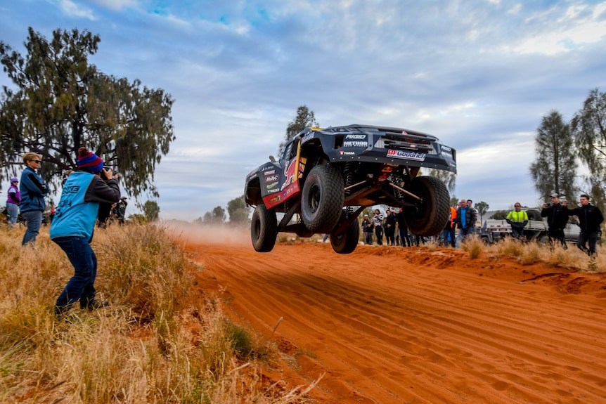 A black four-wheel-drive in the air, about a metre off the ground, with a red-dirt track beneath it and spectators watching on.