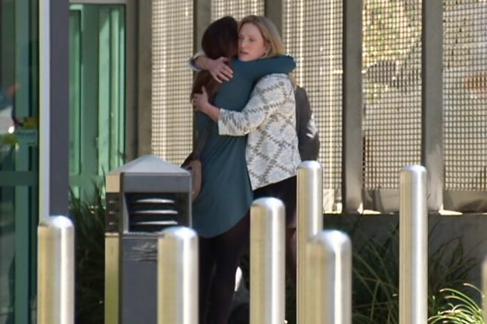 A blonde woman wearing a cardigan hugs another woman outside what looks like a court complex.