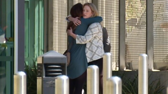 A blonde woman wearing a cardigan hugs another woman outside what looks like a court complex.